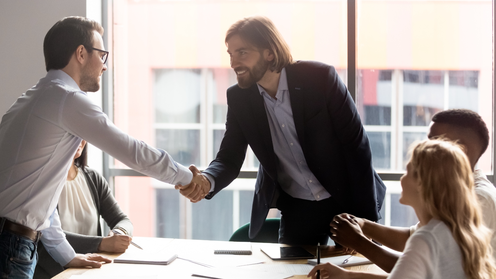 Two people shake hands at a table surrounded by other people.