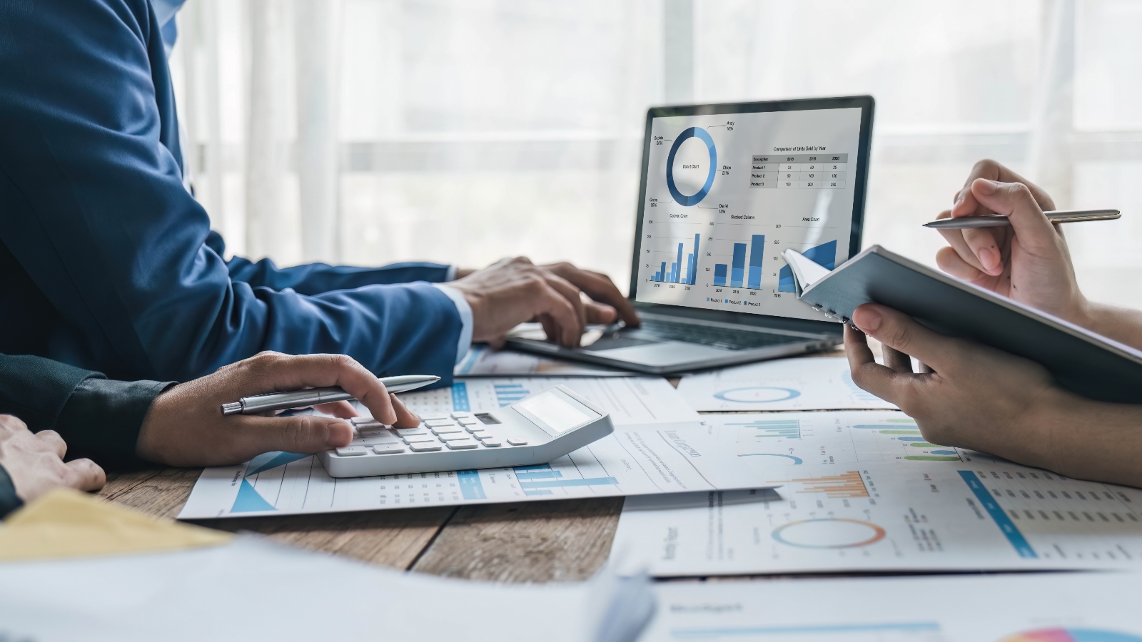 Three accountants compare financial data with charts across paper and laptops.